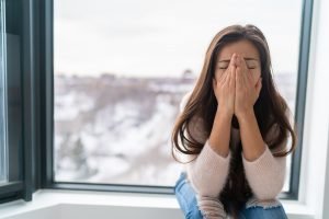 unhappy woman sits on a window ledge with her hands over her face. She gets help from CBT cognitive behavioral therapy in Burke, VA from Nova Terra therapy